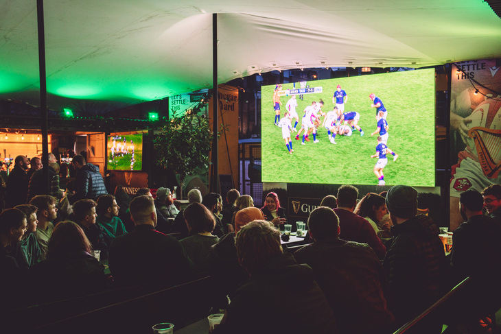People watching rugby on a screen screen, beneath an outdoor covering