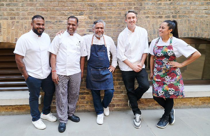 Five chefs wearing chef's whites and aprons, posing against a brick wall